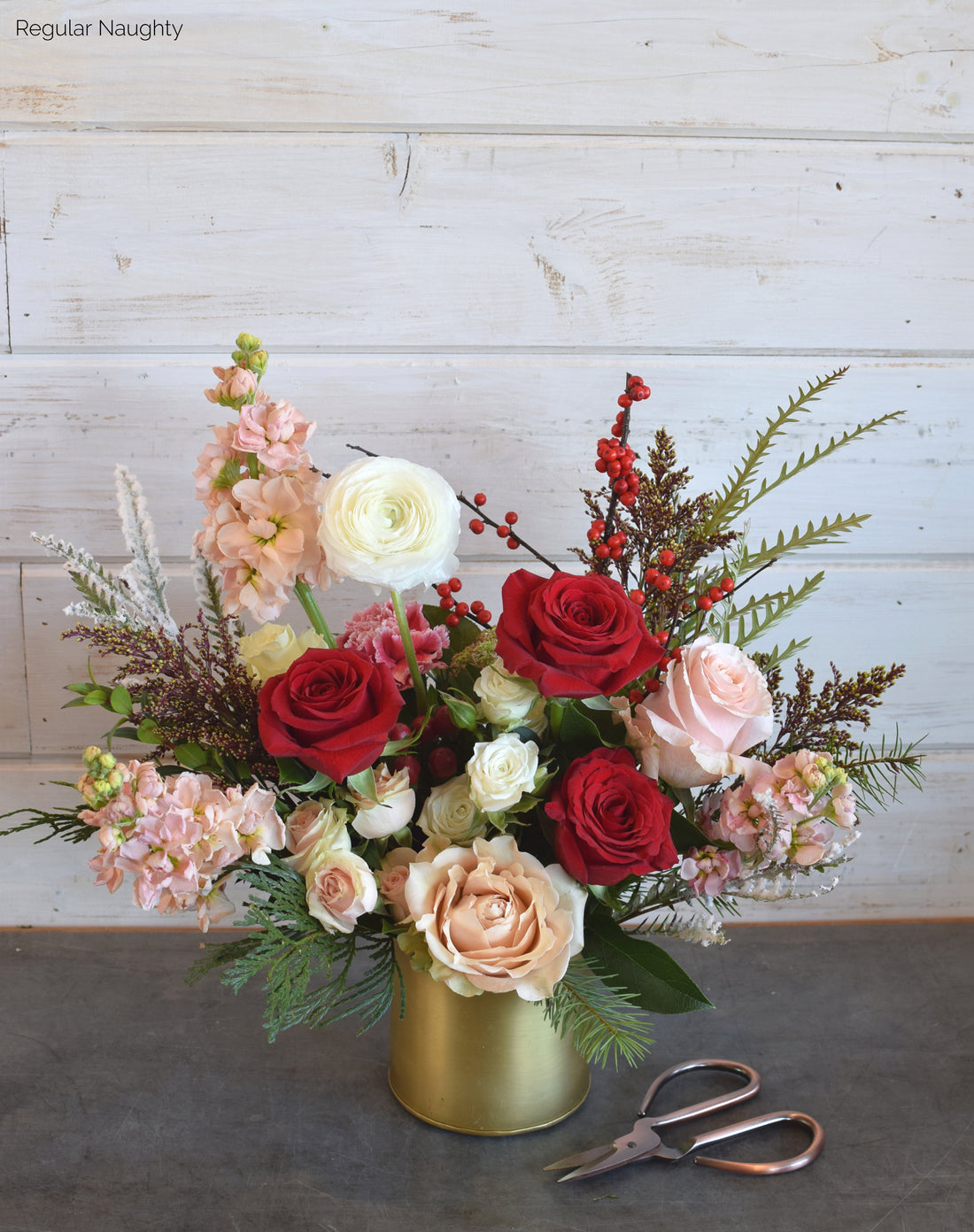 Naughty colorway of the Sleigh All Day holiday flower arrangement featuring bold red roses, ranunculus, and ilex berries, styled with winter greens in a chic gold vessel.