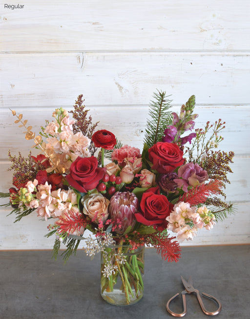 Vibrant Naughty colorway Christmas bouquet featuring bold red roses, spray roses, garden roses, fragrant stock, and festive pine, accented with coffee bean, button daisies, and carnations, arranged in a modern glass vase for a festive holiday touch.