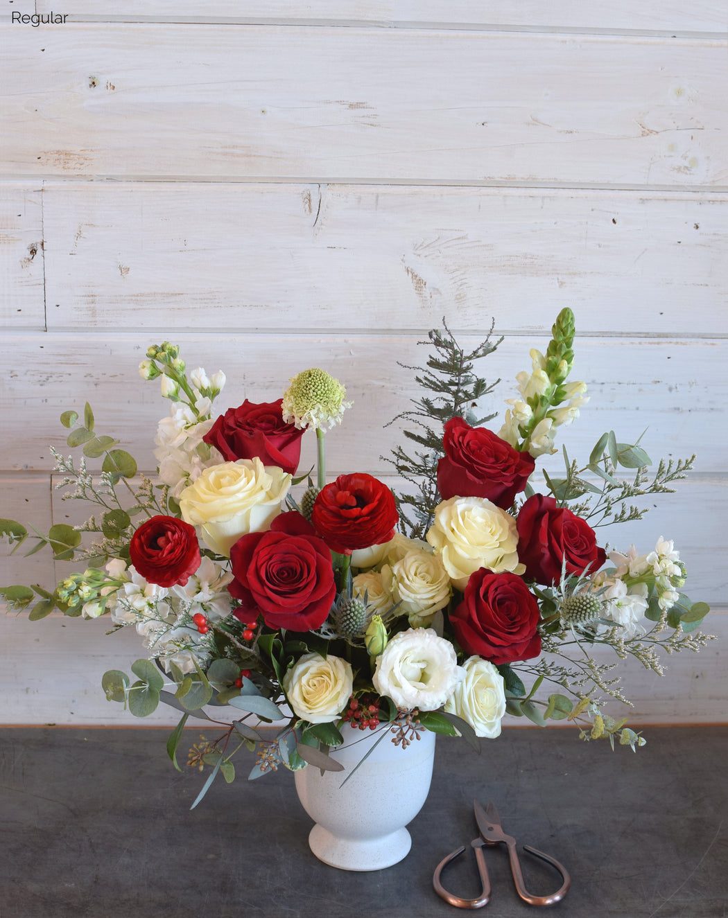 The Keeper flower arrangement featuring red and white roses, ranunculus, lisianthus, snapdragon, scabiosa, eucalyptus, and textured foliage in a sleek white compote vase.