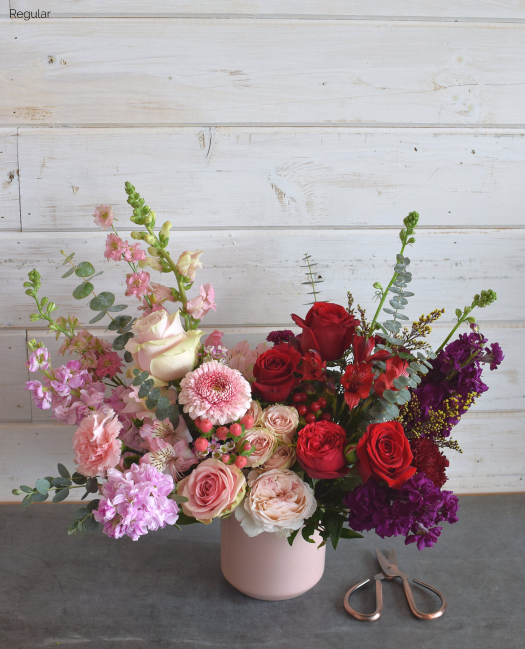 A Valentine ombre floral bouquet from pink to red and dark purple with a variety of flowers including roses and daisies, arranged in a pink ceramic vase.