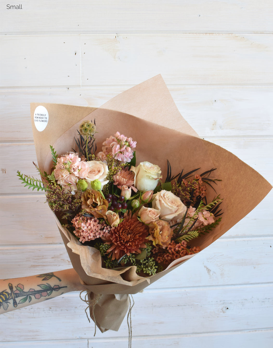 A warm-toned bouquet featuring roses, spray roses, chrysanthemums, lisianthus, coffee bean stock, button daisies, brown solidago, and beige and brown foliage, wrapped in kraft paper and tied with twine.