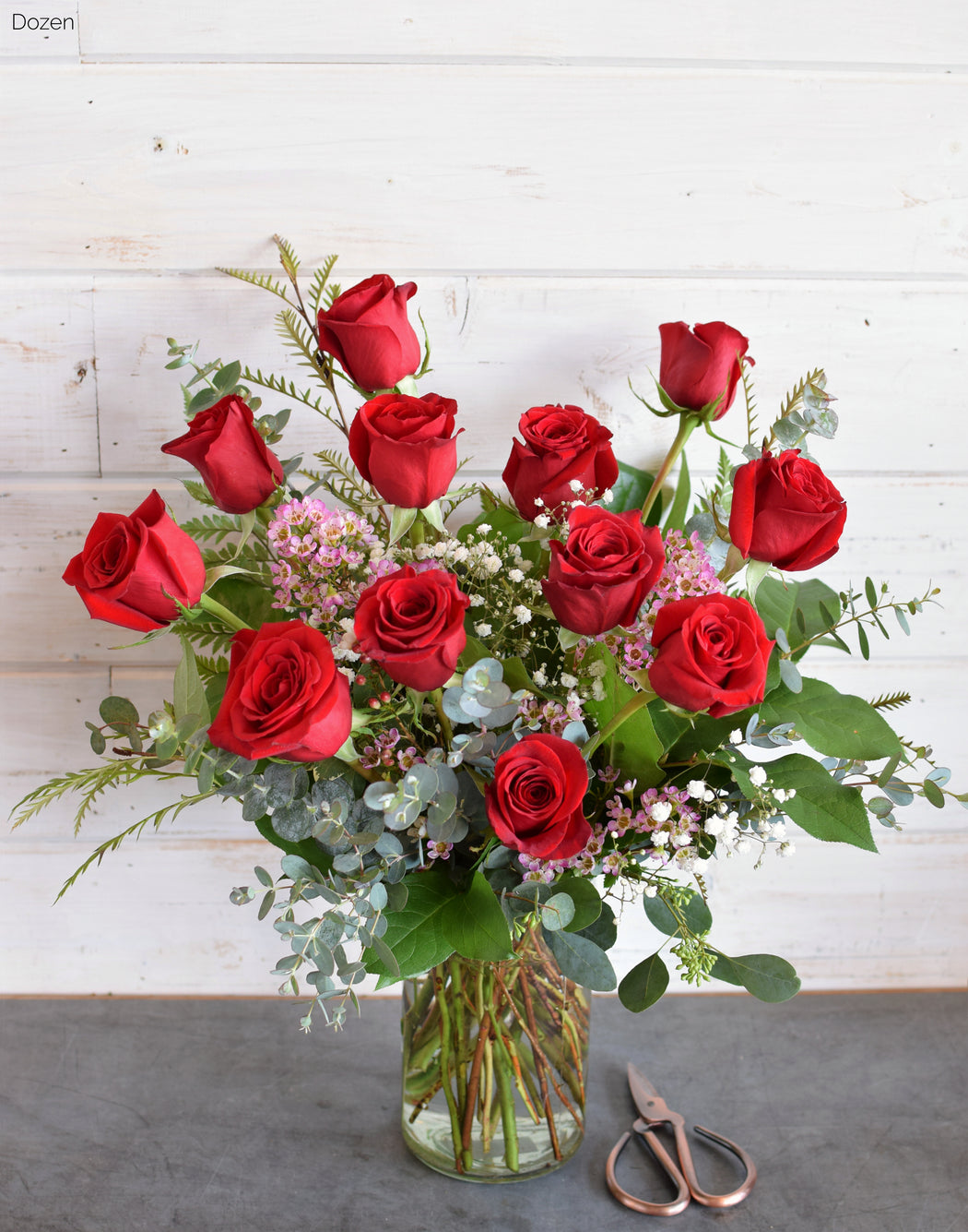 A dozen red roses arranged in a clear glass vase, with baby's breath and greenery for embellishment, displayed on a white surface.