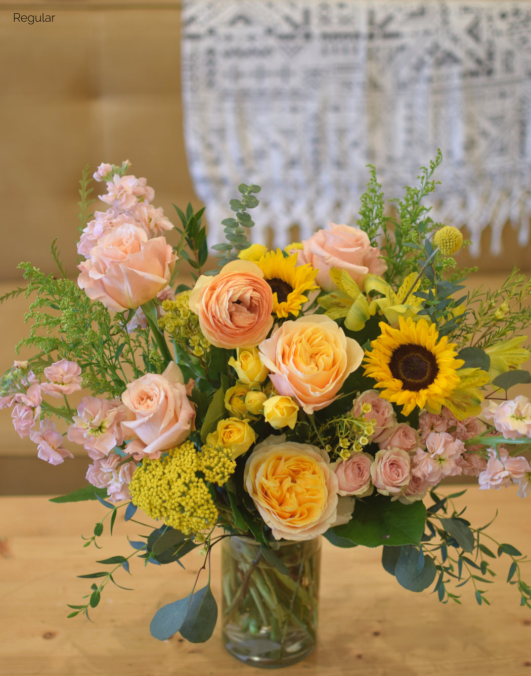 Bouquet of flowers with yellow sunflowers and peach roses called the Custom  Vibe regular size in a clear vase on a wooden surface.