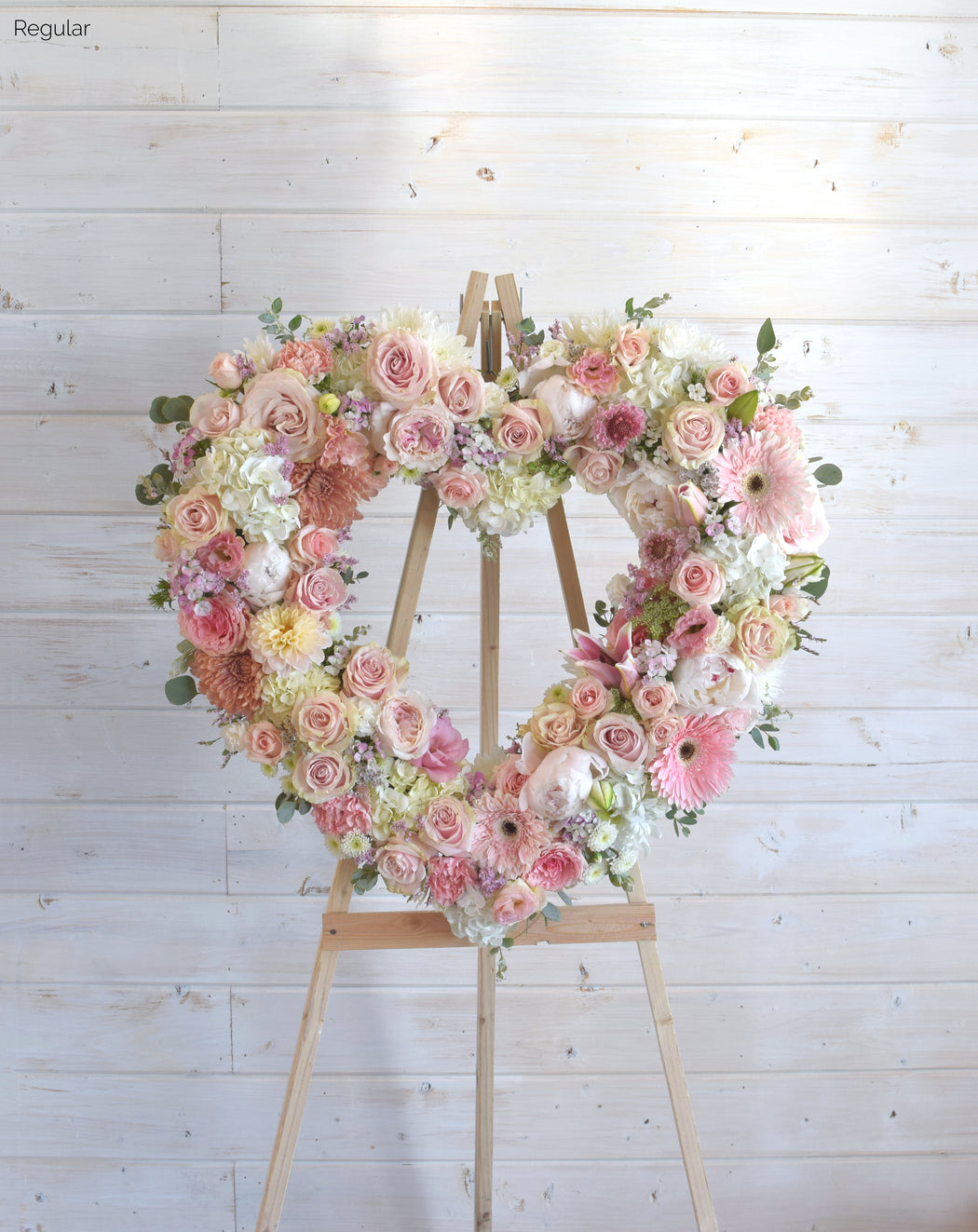 Regular-sized Open Heart wreath in soft pink and white hues, featuring roses, peonies, dahlias, spray roses, hydrangea, and fresh greenery, displayed on an easel for a heartfelt tribute.