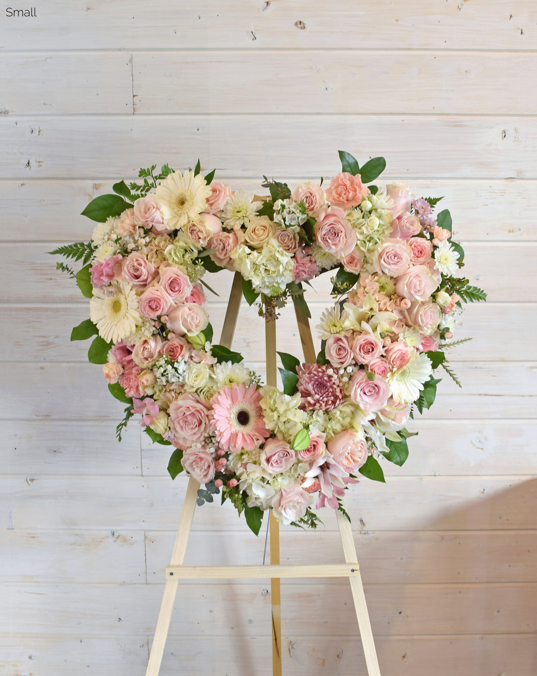 Regular-sized Open Heart wreath in pink and white, featuring roses, spray roses, hydrangea, lilies, and fresh greenery, displayed on an easel for a heartfelt sympathy tribute."