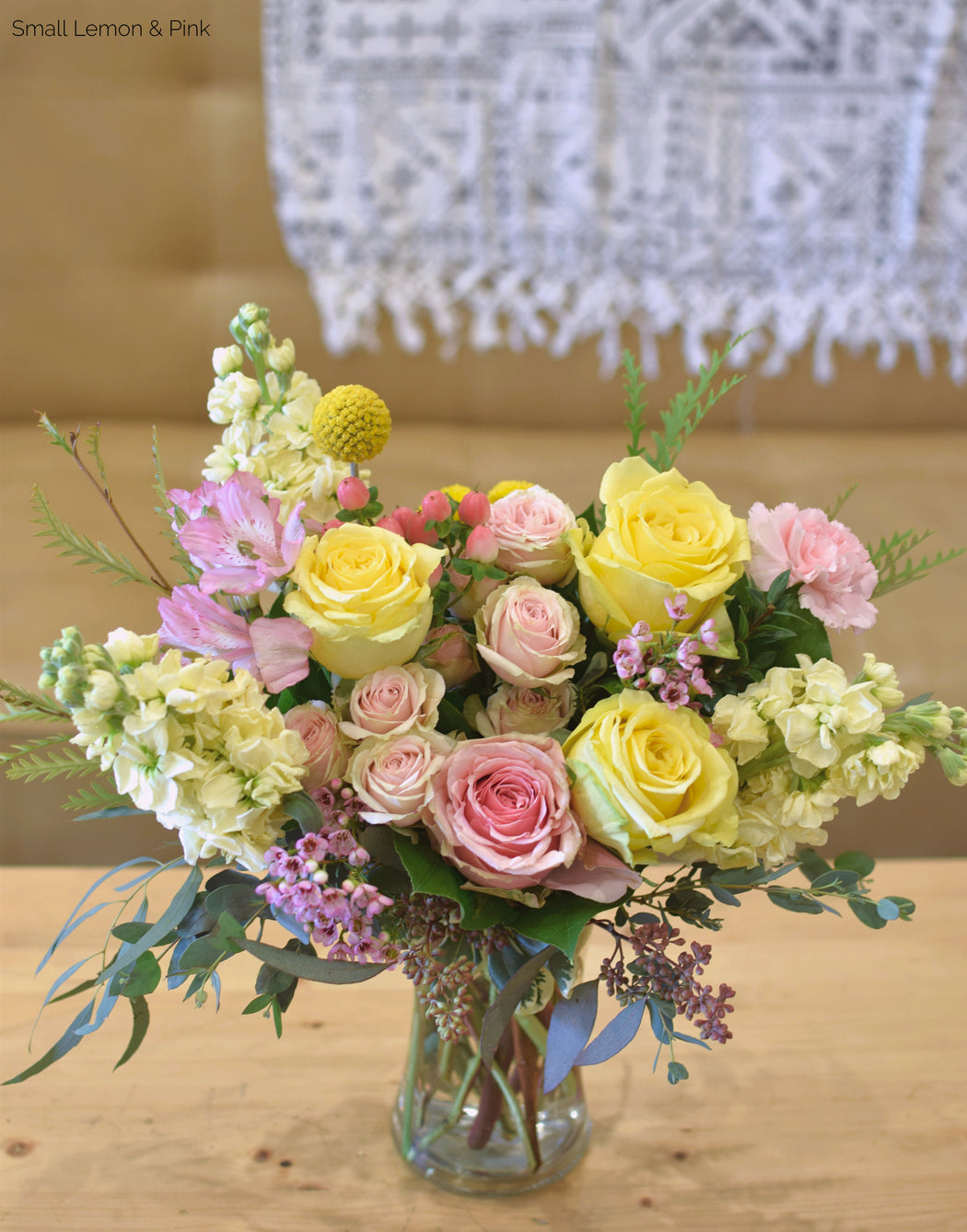 Our small-sized bouquet of pastel flowers, called the Custom Vibe in a clear vase with pink and lemon colored blooms on a wooden surface with a blurred background.