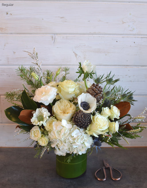 The Noel winter bouquet featuring white Mondial roses, hydrangea, anemone, pine cones, blue thistle, magnolia leaves, and winter foliage in a sleek glass vase.