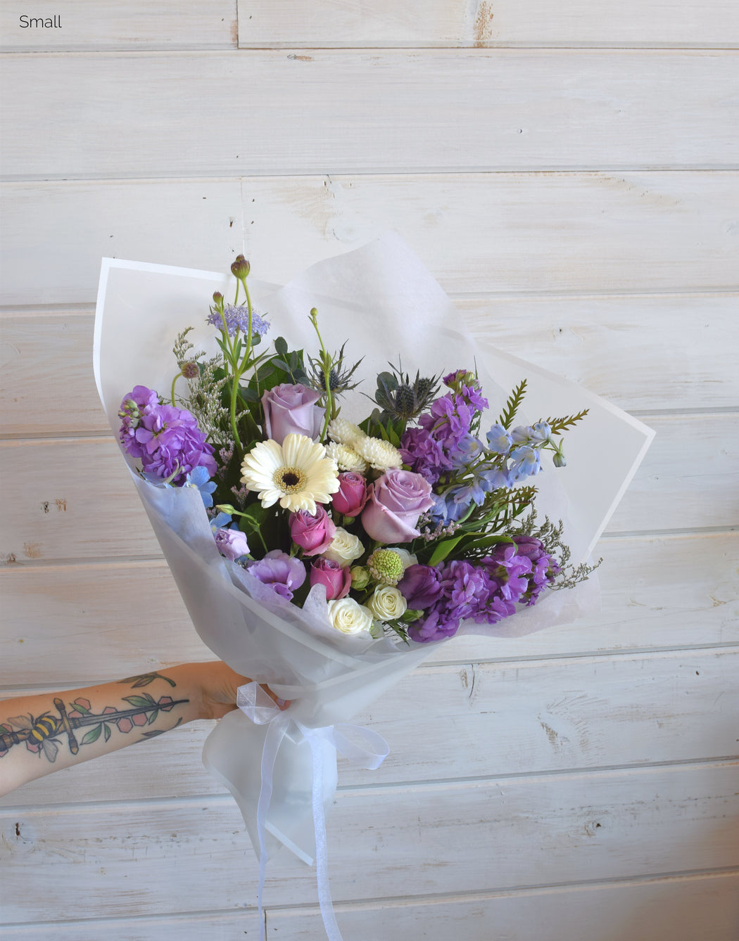 Small Wild Berry Wrap bouquet with roses, spray roses, tulips, lisianthus, blue delphinium, eucalyptus, and filler, wrapped in white paper with satin ribbon.