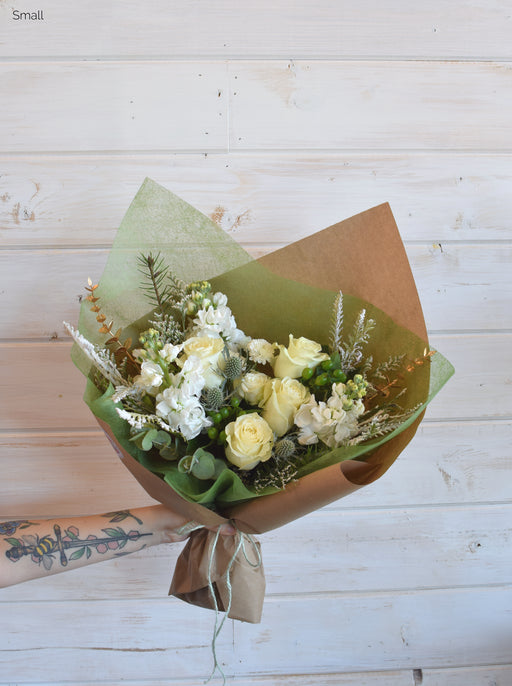 A festive holiday bouquet featuring crisp white roses, winter greens, and gold accents, wrapped in Kraft paper and tied with holiday twine.