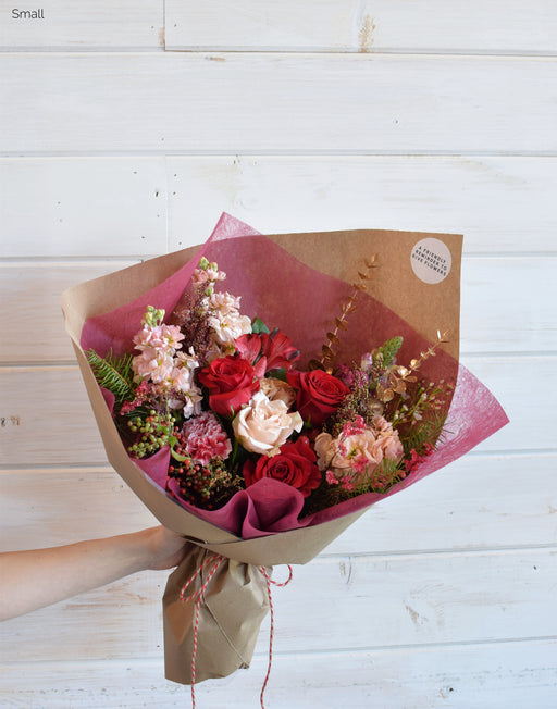 A bold holiday bouquet featuring red roses, warm peaches, snapdragon, alstroemeria, and winter greens, wrapped in Kraft paper with festive twine.